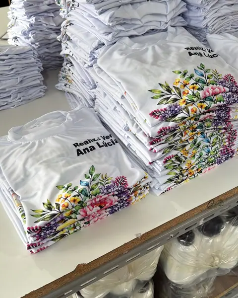 Pilha de camisetas brancas com flores coloridas e o nome Ana Lúcia